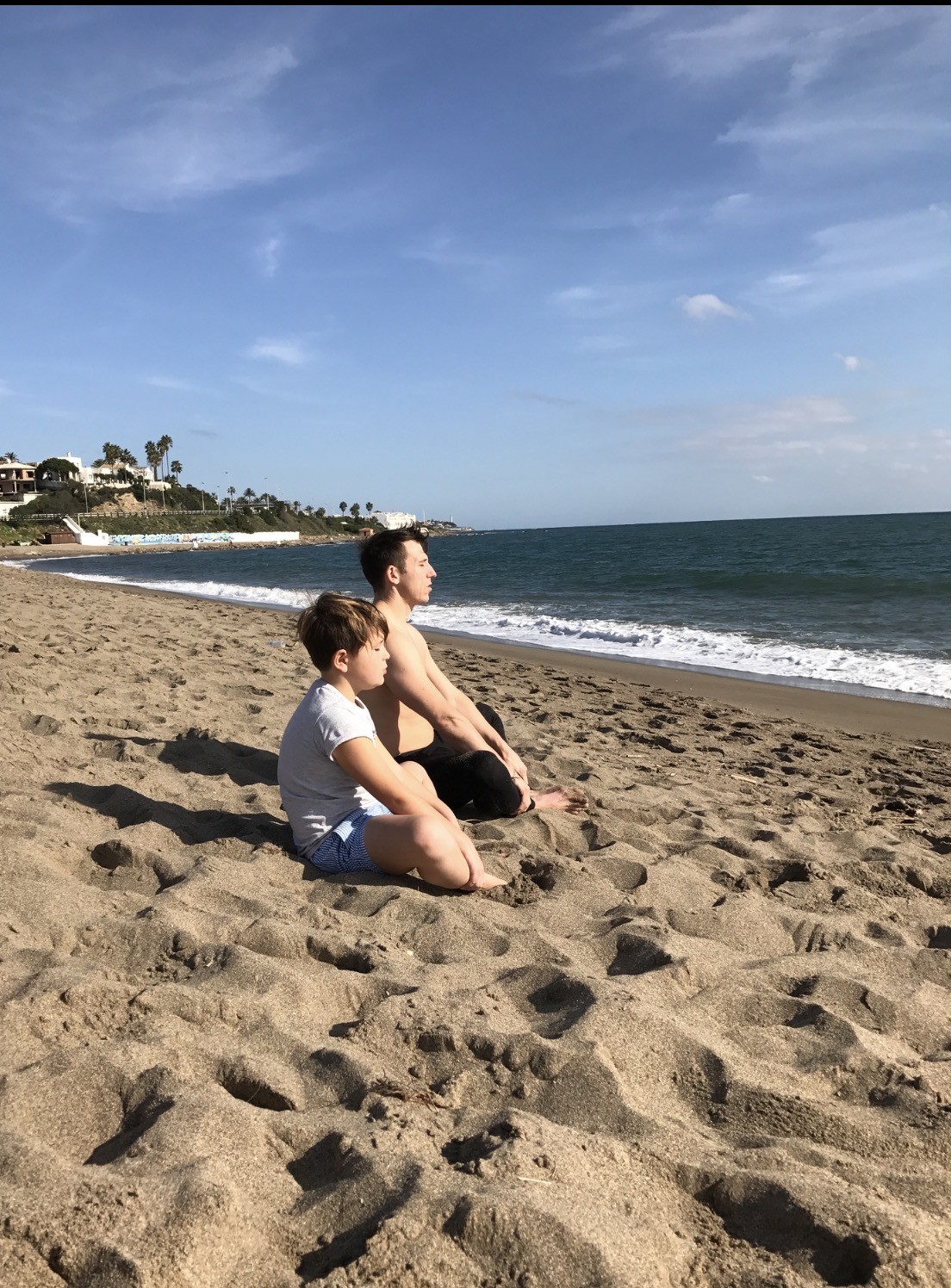 Gary and his son Thomas sitting cross-legged on a beach, eyes closed, breathing