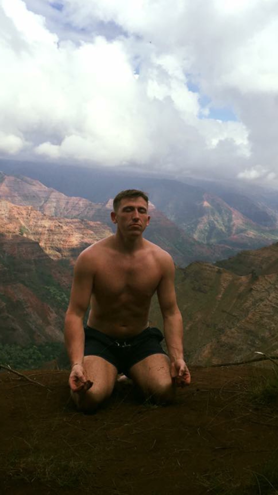 Gary Andrew Rhodes kneeling on a canyon ridge, eyes closed, under a cloudy sky